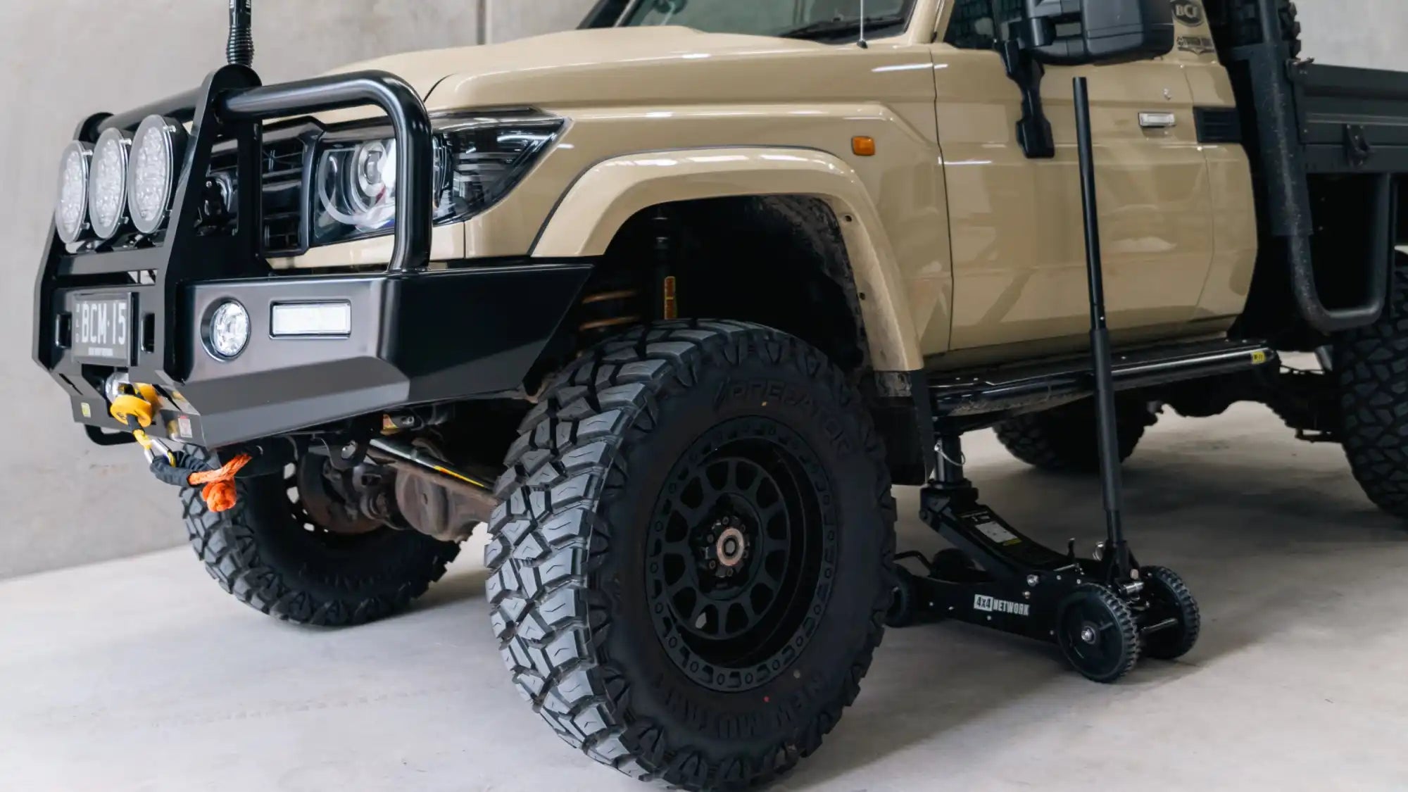 Lifted tan off-road truck with a front bull bar, auxiliary lights, and a floor jack.